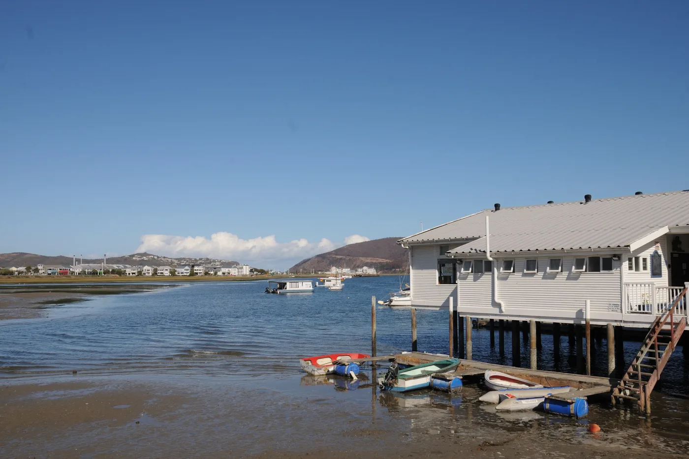 Boats and a timber boathouse on stilts over the Knysna lagoon at low tide, with hills behind